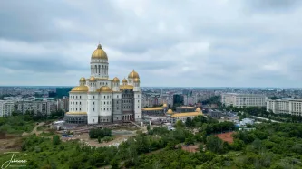 An elevated view showcases the monumental People's Salvation Cathedral (Catedrala Mântuirii Neamului) in Bucharest, Romania. This massive Eastern Orthodox structure, still undergoing exterior work, is designed to be the tallest domed church in the world, symbolizing national unity and faith. Its towering central dome and multiple surrounding domes are covered in brilliant gold, making it a radiant landmark visible across the urban landscape. Surrounded by green spaces on the Dealul Arsenalului (Arsenal Hill), this grand project contrasts sharply with the density of the surrounding city and stands as a powerful new focal point in the Romanian capital.