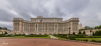 An awe-inspiring ground-level panorama of the Palace of the Parliament in Bucharest, Romania. This gargantuan edifice, also known as the People's House (Casa Poporului), stands as one of the world's largest administrative buildings and a lasting symbol of communist-era megalomania. Its immense neoclassical façade, stretching seemingly endlessly across the frame, features countless arched windows and intricate detailing. The Romanian flag proudly flies before its central entrance, while the golden domes of the People's Salvation Cathedral are just visible, hinting at the city's evolving skyline, under a dramatic, overcast sky.