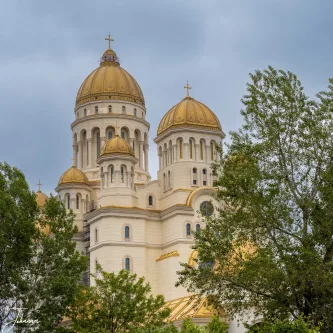 A majestic close-up reveals the stunning People's Salvation Cathedral (Catedrala Mântuirii Neamului) in Bucharest, Romania. This grand Orthodox cathedral, which is still undergoing final completion phases, stands as a profound symbol of faith and national identity. The image beautifully highlights its most striking features: the multiple, resplendent golden domes and intricate Byzantine-inspired architecture, all crowned with crosses reaching towards the heavens. Framed by lush green trees and set against a dramatic, overcast sky, its radiant presence creates a powerful visual contrast, signifying a new chapter in the city's spiritual landscape.