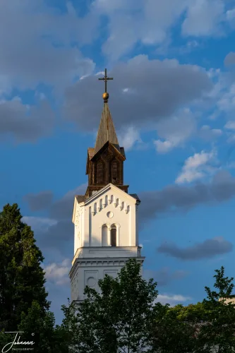 Reaching for the dramatic sky, this elegant church tower captures the last golden light of day. Its pristine white facade and prominent dark spire stand as a symbol of enduring faith, beautifully framed by the deep blue and cloud-strewn heavens. A beacon in the changing light.