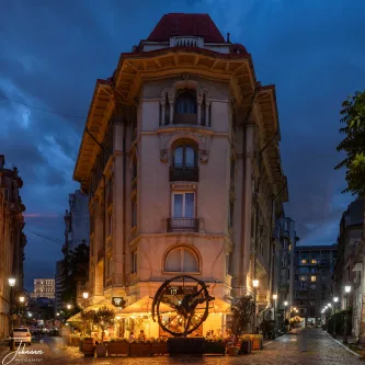 The golden glow of a cafe spills onto the cobblestones, warming the dramatic, twilight sky. This historic street corner in Bucharest captures the city's perfect blend of architectural drama and cozy evening life.